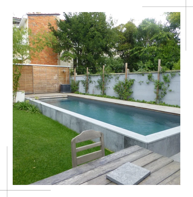 A modern rectangular swimming pool surrounded by a concrete edge and lush green grass, enhanced by wooden fencing with climbing plants lining the poolside. A pergola nearby shelters a wooden table and chair in the foreground, while leafy trees frame the backdrop beautifully.