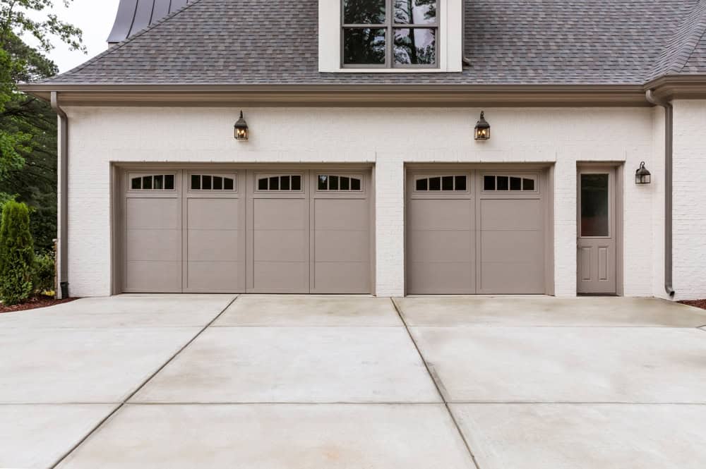 The house exterior features three beige garage doors and a side door set in a light brick facade. The concrete driveway leads up to it, accentuated by a dark gray roof. Two black lantern-style lights perch above the garage doors, while trees grace the background, hinting at potential for fire pits nearby.