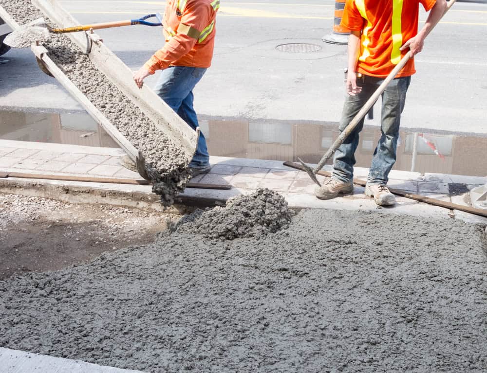 Two construction workers in orange safety vests pour and spread wet concrete onto a sidewalk using a chute and a rake. They are working near an outdoor kitchen builder's site on a sunny day, right by the street curb.