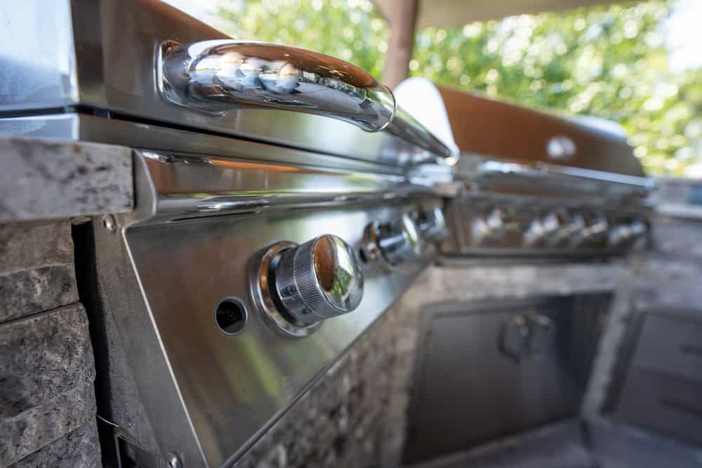Close-up of a modern outdoor stainless steel grill with shiny knobs and a stone countertop, set beneath a stylish pergola. The background features green foliage, partially blurred.