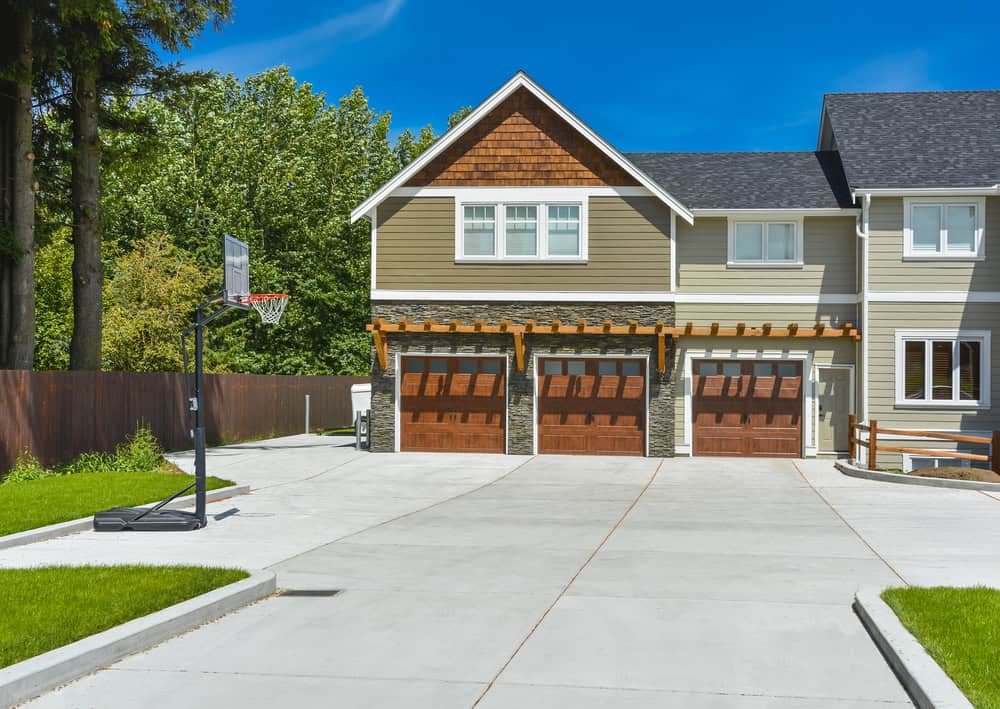 A suburban house with a three-car garage featuring wooden doors and a basketball hoop on the wide concrete driveway. The beige siding, white trim, and gabled roof complement the lush trees in the background. A cozy fire pit adds warmth and charm to this inviting home setting.
