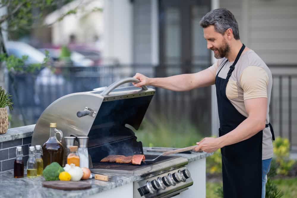 A man wearing an apron grills meat on a gas barbecue next to a beautifully designed pergola in an outdoor setting. Various ingredients like vegetables, oil, and seasonings are on the counter nearby. He smiles as he flips the meat with a spatula.