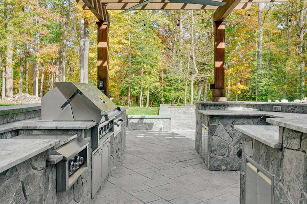 Outdoor kitchen with stone countertops and a stainless steel grill under a pergola, crafted by expert outdoor kitchen builders. The area is surrounded by lush trees with autumn foliage, creating a cozy, natural setting.