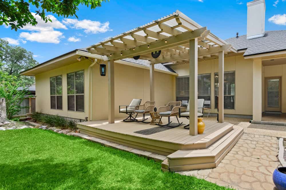 An outdoor patio with a wooden pergola and seating area is set on a raised deck adjacent to a beige house. Crafted by expert outdoor kitchen builders, the yard features green grass and stone paving. A lamp hangs from the pergola, illuminating the scene under a clear blue sky.