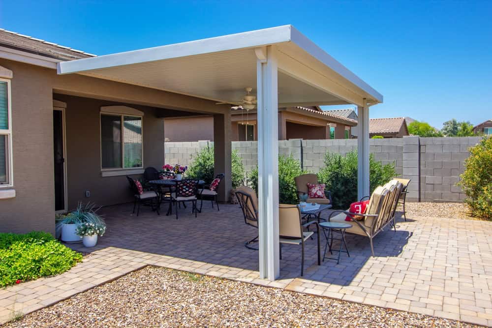 A backyard patio boasts a covered pergola. There's a table with chairs and a cozy seating area around one of the elegant fire pits. Green shrubs border the stone-paved patio, while a stone wall encloses the area. It's a sunny day with a clear blue sky.