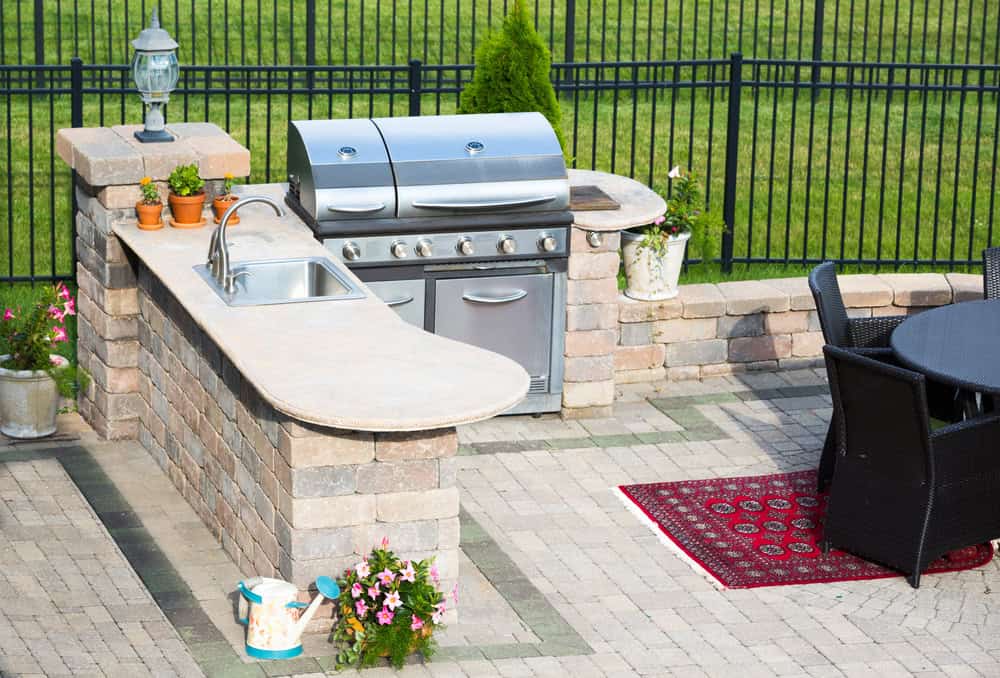 Outdoor kitchen with a stainless steel grill, sink, and stone countertops sits gracefully under charming pergolas. Potted plants and a watering can decorate the area. A woven black dining set and decorative rug are nearby, with a grassy lawn and metal fence in the background.