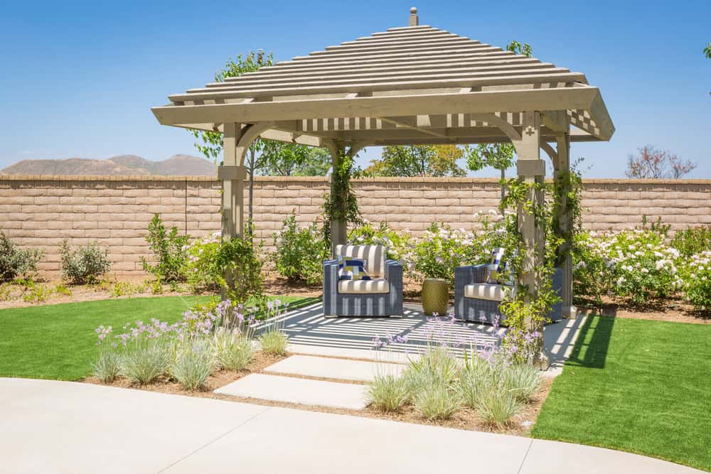 A garden gazebo with a wooden pergola roof stands on a patio. Two cushioned armchairs with striped fabric sit inside. Vines climb the structure, and flowers and green grass surround the area. Nearby, outdoor kitchen builders work against the backdrop of a stone wall under the blue sky.