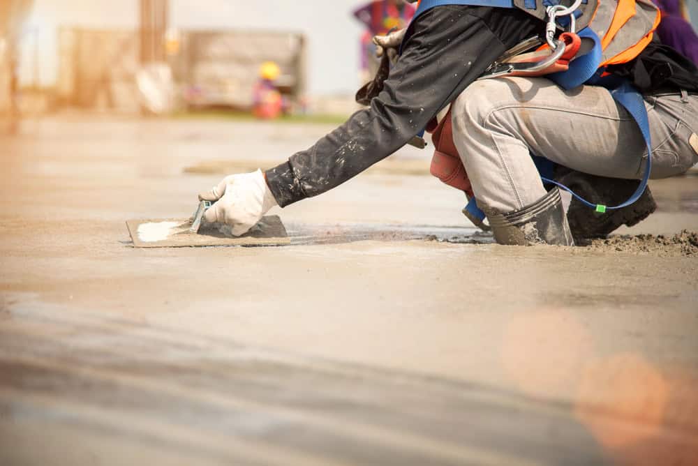 A construction worker kneels on a concrete surface, smoothing it with a trowel as they prepare an outdoor fireplace. They are wearing gloves, a safety harness, and a reflective vest. The sun shines brightly, creating lens flares in the foreground.